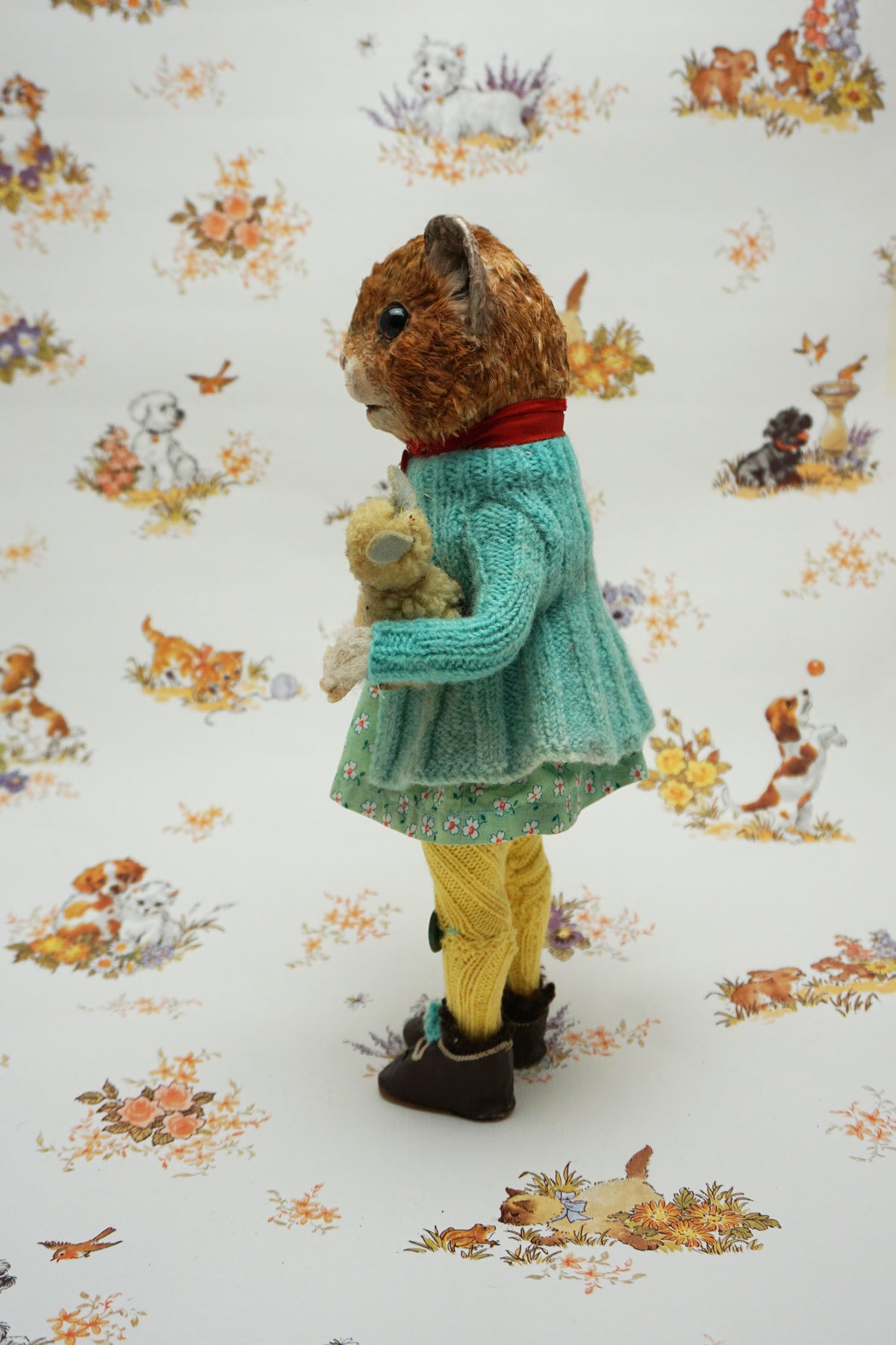 A plush guinea pig dressed in a teal cardigan, floral dress, and yellow tights holds a small stuffed animal. The background features whimsical illustrations of animals and nature scenes.
