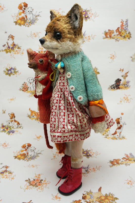 A plush fox toy dressed in a patterned red and white dress, teal cardigan, and red boots holds a smaller red plush creature. The background features a whimsical animal print with various small animals.