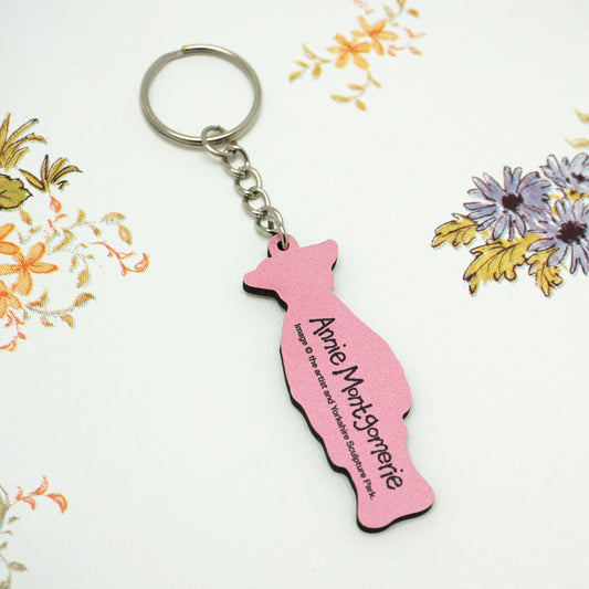 The back of the Wilton keyring is pink with the text "Annie Montgomerie" printed on it. The keyring is placed on a white surface with a floral pattern featuring orange and purple flowers.