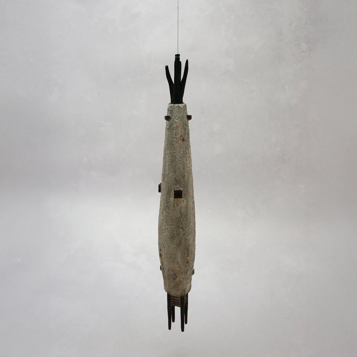 Hanging wooden house in a long slender shape with four legs at the bottom. The house is grey with dark brown windows and turrets and it has grey thread from the top which is hanging the piece