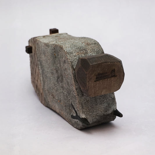 A grey hand carved wooden house with two small legs and a boulder holding it up. The roof is textured and had a small chimney and the house has two small wooden windows.