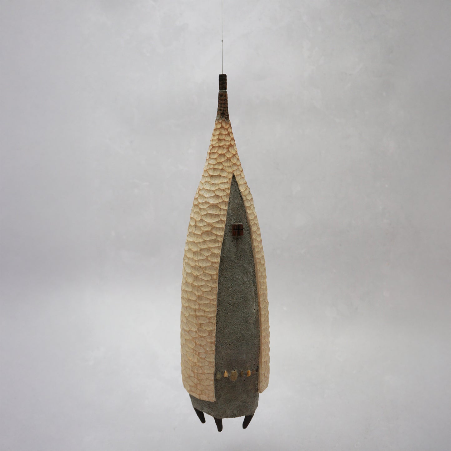 A floating wooden house featuring a hand carved pattern on the sides and back, and a grey mud like colour on the front. The house has four feet and a carved wooden window at the front. The house is hanging from grey thread.