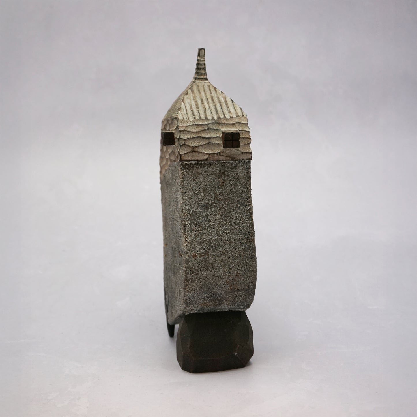 A grey hand carved wooden house with two small legs and a boulder holding it up. The roof is textured and had a small chimney and the house has two small wooden windows.