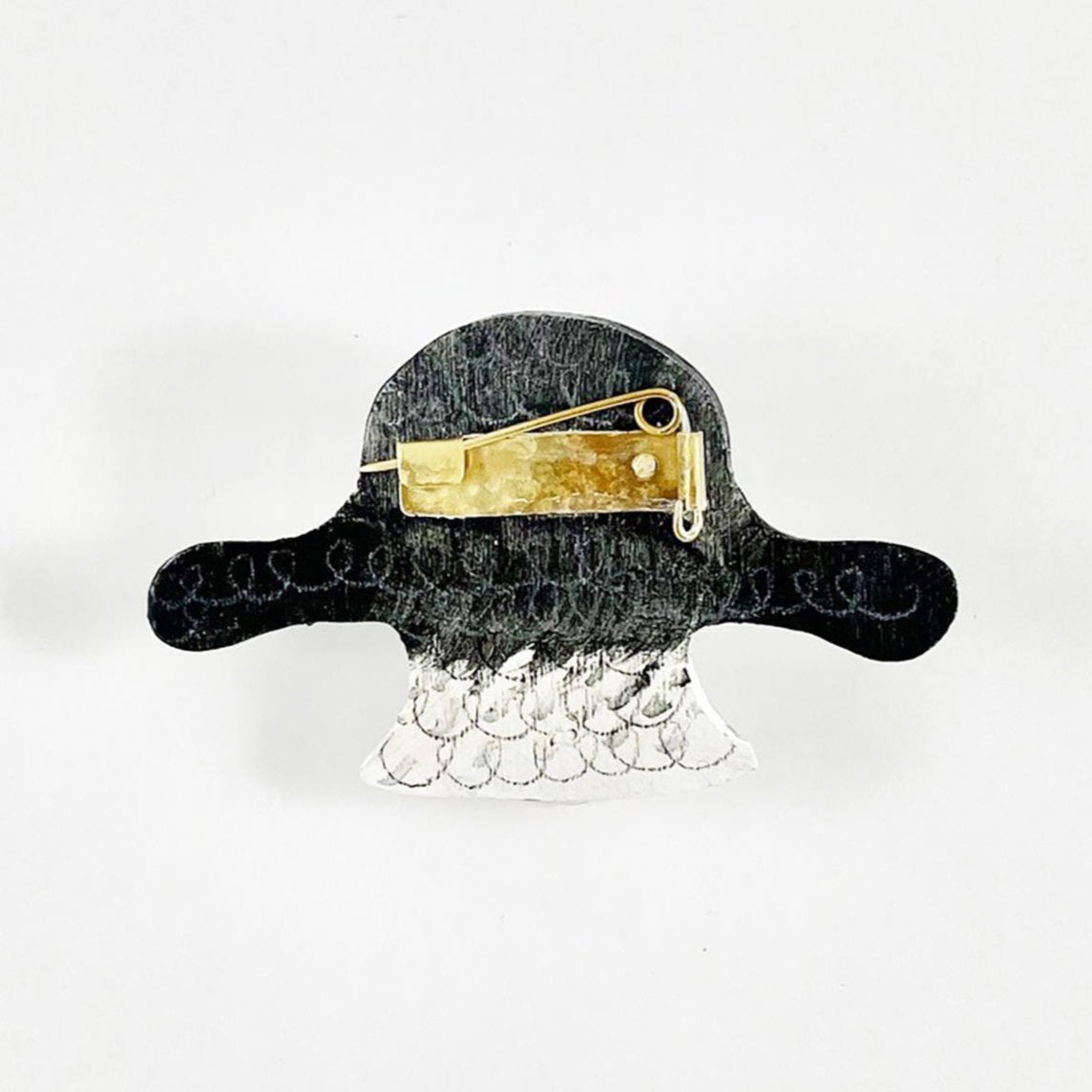 The back of a quirky brooch shaped like a goat's head, featuring a black top and a white base. A gold pin is attached, adding a playful touch to the design. The background is plain white, highlighting the brooch's details.