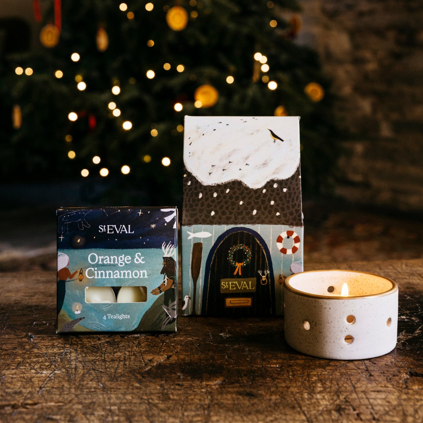St Eval paper cottage packaging featuring a snowy roof and brown door, next to a small box of tealights and a ceramic candle holder.