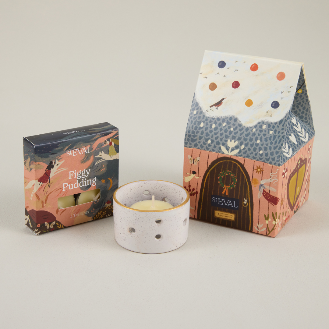 St Eval paper cottage packaging featuring a snowy roof and brown door, next to a small box of tealights and a ceramic candle holder.