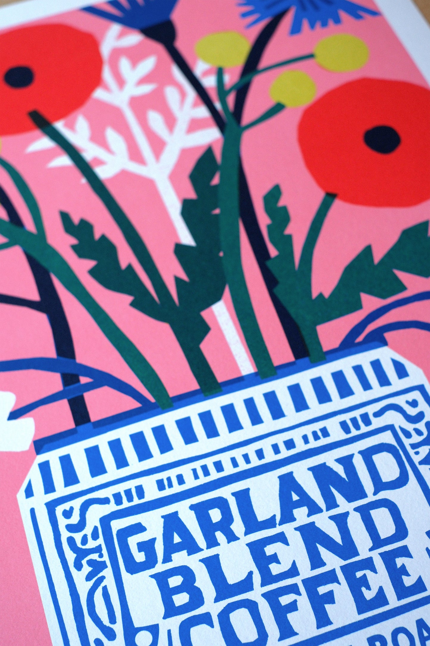 A vibrant illustration featuring a blue and white coffee canister labelled "Garland Blend Coffee" surrounded by a bouquet of colourful flowers, including red poppies and blue blooms, set against a pink background.