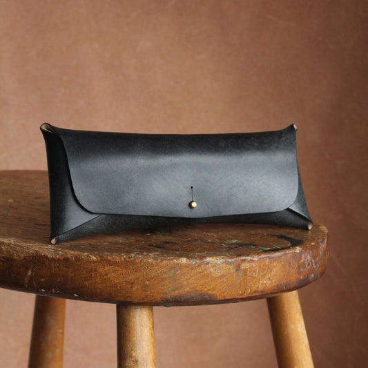 A smooth and simple black leather glasses case with a brass stud closure, placed on a wooden stool.
