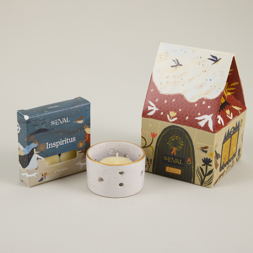 St Eval paper cottage packaging featuring a snowy roof and brown door, next to a small box of tealights and a ceramic candle holder.