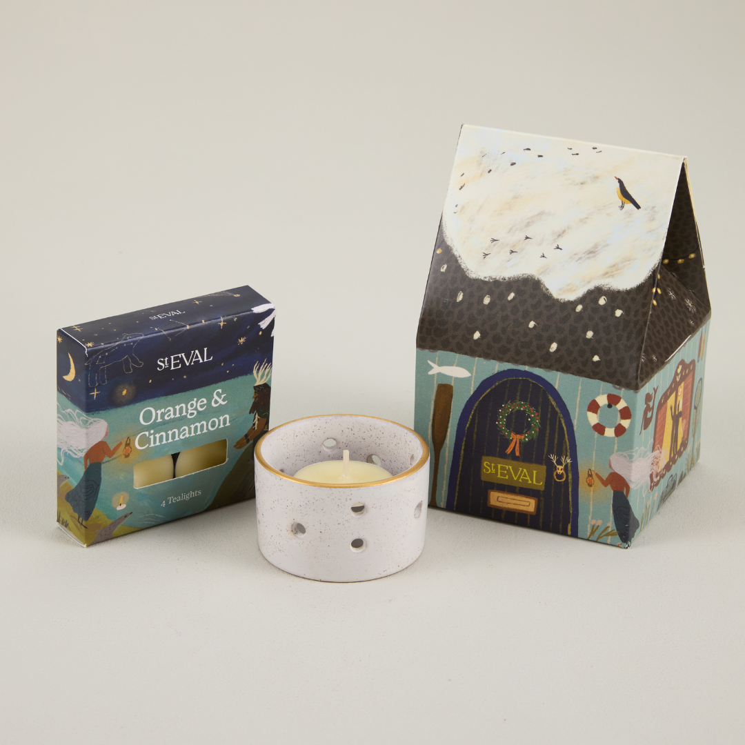 St Eval paper cottage packaging featuring a snowy roof and brown door, next to a small box of tealights and a ceramic candle holder.