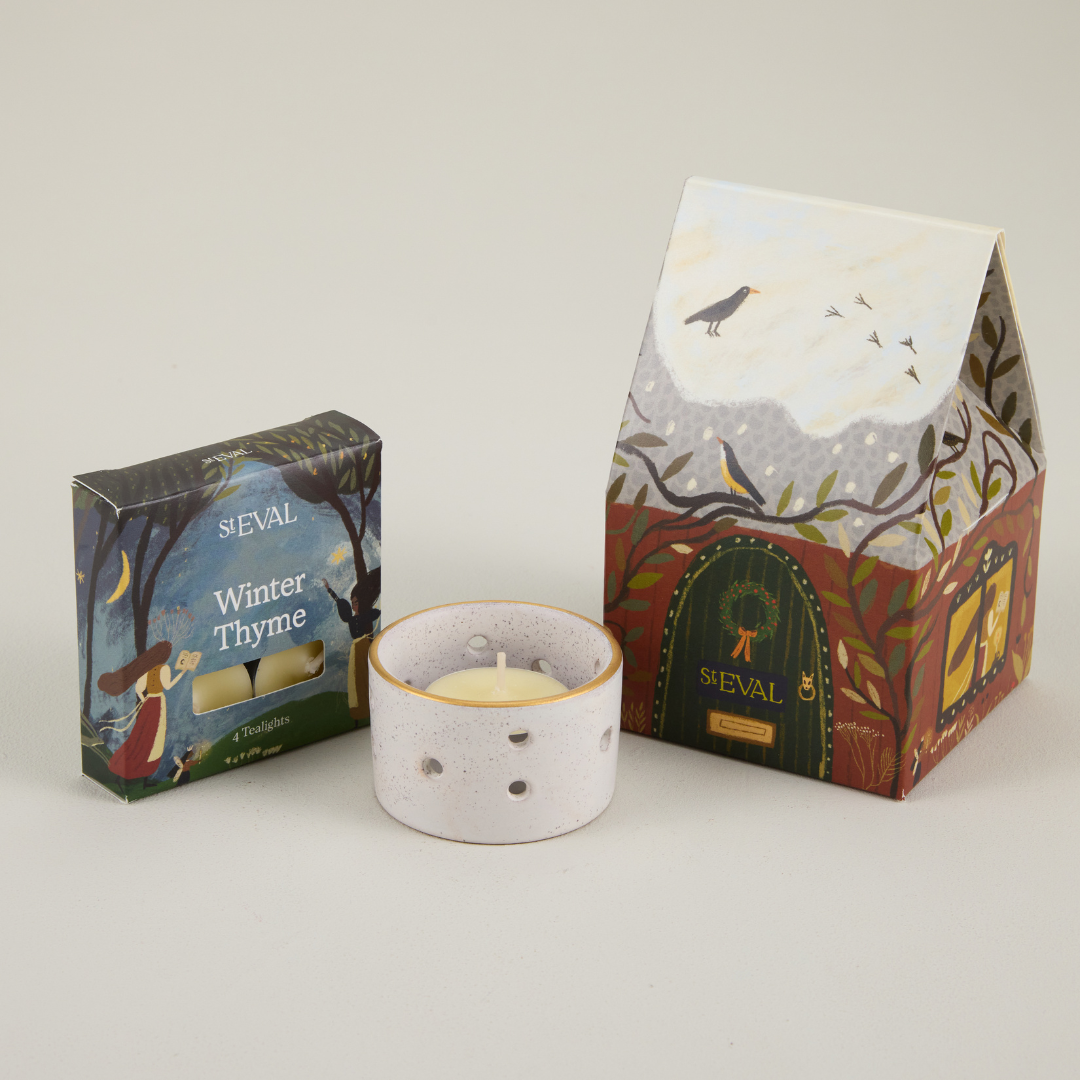 St Eval paper cottage packaging featuring a snowy roof and brown door, next to a small box of tealights and a ceramic candle holder.