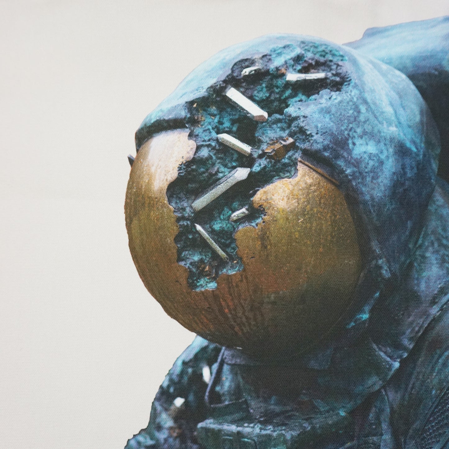 A close-up of a weathered bronze astronaut sculpture, featuring a damaged helmet with a partially exposed golden sphere. The surface shows signs of corrosion and wear, highlighting the intricate details of the figure's suit and the surrounding texture.