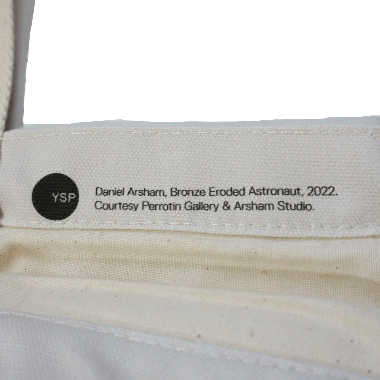 A close-up of a label attached to a fabric item, featuring the text: "Daniel Arsham, Bronze Eroded Astronaut, 2022. Courtesy Perrotin Gallery & Arsham Studio." The label includes a circular logo marked "YSP.