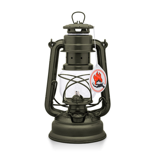 A vintage-style oil lantern in a dark green finish. It features a glass reservoir, a protective wire cage, and a hanging handle. A tag with a flame logo is attached, indicating it is a new product. The lantern is set against a plain white background.