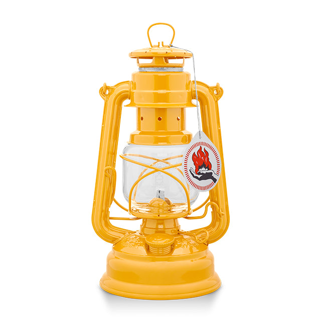 A vintage-style oil lantern in a dark yellow finish. It features a glass reservoir, a protective wire cage, and a hanging handle. A tag with a flame logo is attached, indicating it is a new product. The lantern is set against a plain white background.