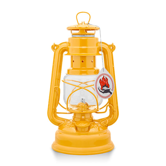 A vintage-style oil lantern in a dark yellow finish. It features a glass reservoir, a protective wire cage, and a hanging handle. A tag with a flame logo is attached, indicating it is a new product. The lantern is set against a plain white background.