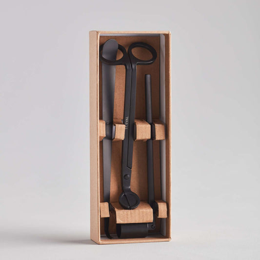 A neatly packaged set of black scissors and tools, displayed in a cardboard box with compartments. The tools include a pair of scissors, a straight tool, and a curved tool, all secured with cardboard inserts. The background is plain and light.