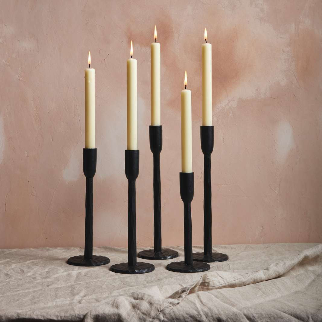 Five tall, slender candles with cream-coloured wax are lit and placed in black candle holders. They stand on a light, textured fabric against a soft, peach-toned wall, creating a warm and inviting atmosphere.