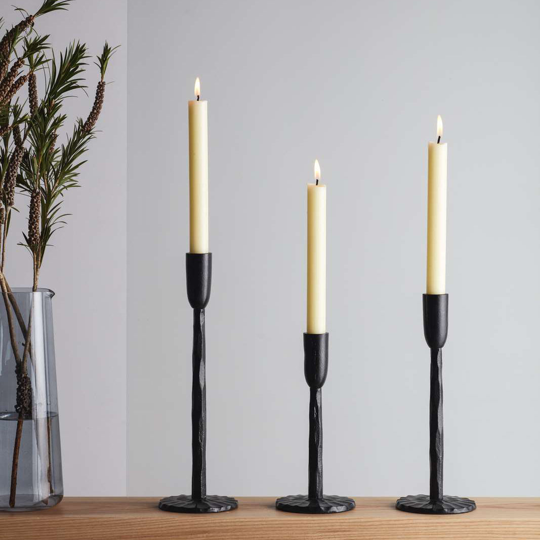 Three elegant candle holders with tall, slender white candles, set against a light grey wall. The holders have a textured black finish and are arranged on a wooden surface. A glass vase with greenery is visible in the background.