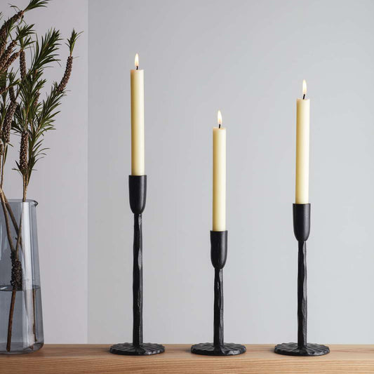 Three elegant candle holders with tall, slender white candles, set against a light grey wall. The holders have a textured black finish and are arranged on a wooden surface. A glass vase with greenery is visible in the background.