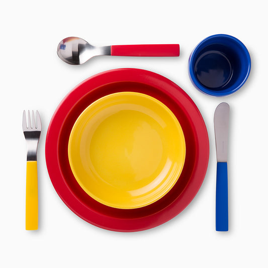 A vibrant table setting featuring a red plate, a yellow bowl, a blue cup, and stainless steel cutlery. The fork and knife have yellow and blue handles, respectively, creating a cheerful and playful dining arrangement.