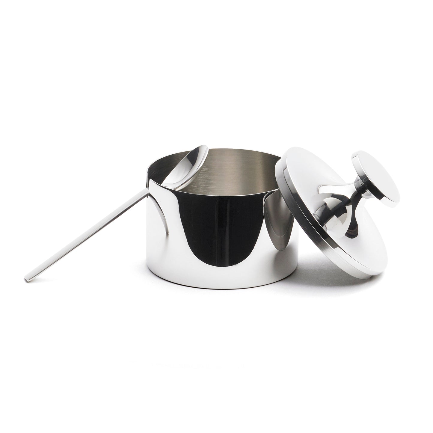 A sleek, stainless steel sugar bowl with a matching lid and a long-handled spoon rests beside it. The bowl has a polished finish, reflecting light, and features a simple, modern design. The lid is slightly ajar, revealing the interior.