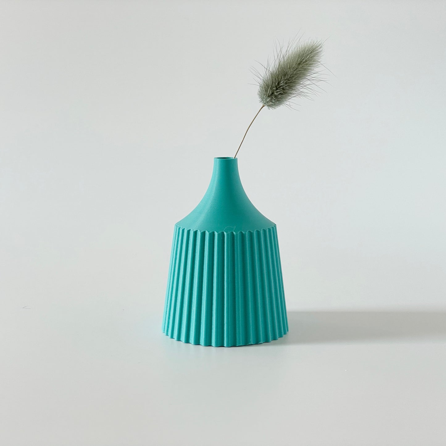 A turquoise, 3D printed vase with a ribbed design, containing a single dried plant.