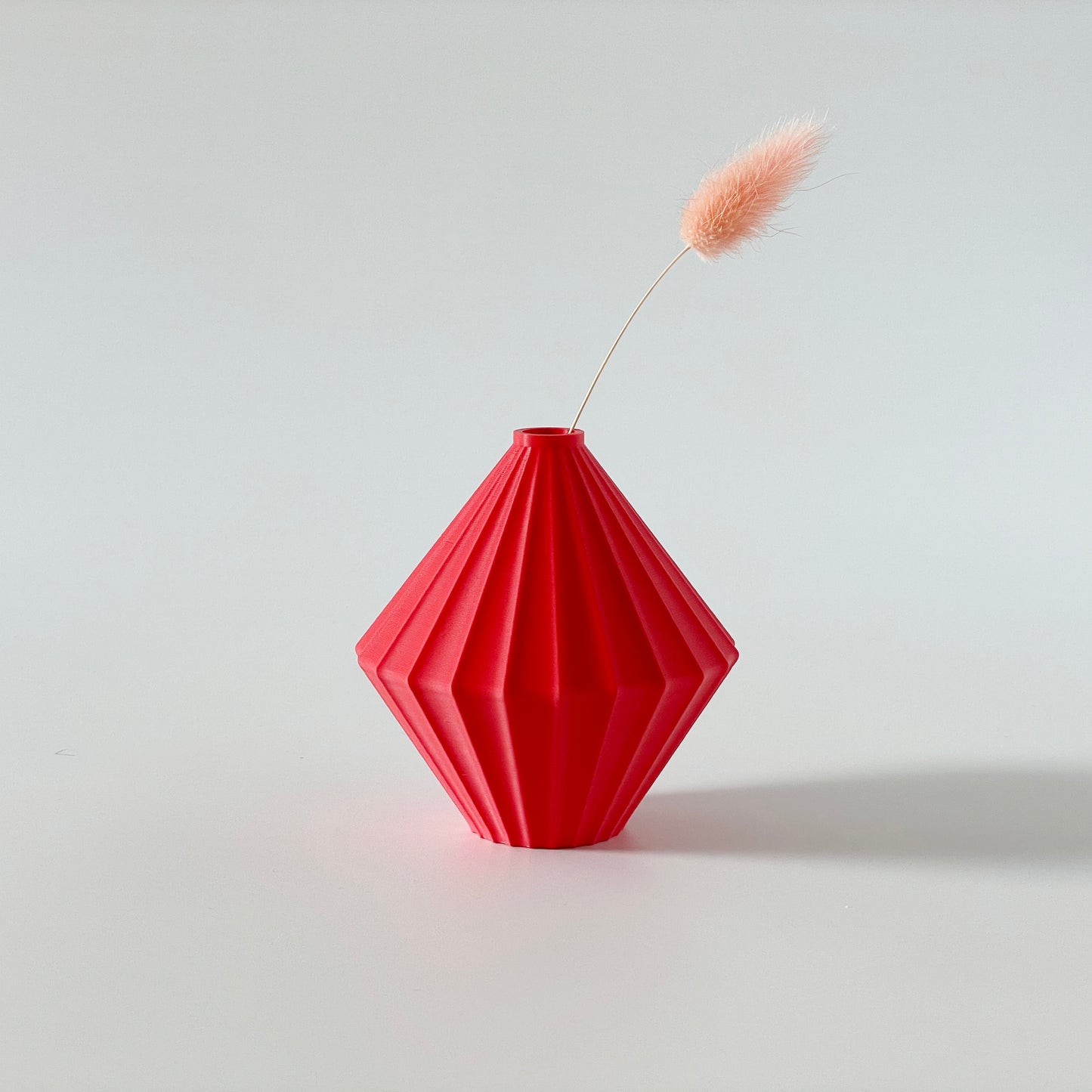 A red, 3D printed triangular vase with a ribbed texture and a small sprig of dried grasses inside on a white background.