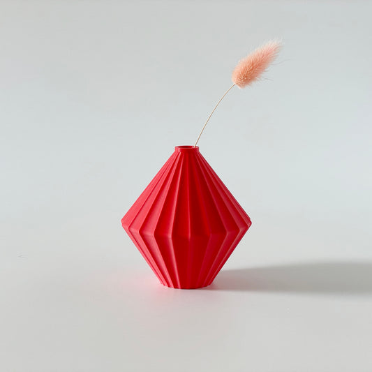 A red, 3D printed triangular vase with a ribbed texture and a small sprig of dried grasses inside on a white background.