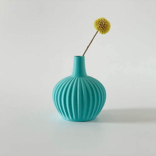 A spherical turquoise, 3D printed vase with a ribbed design, containing a single dried plant.
