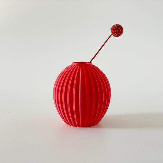 A red, 3D printed spherical vase with a ribbed texture and a small sprig of dried grasses inside on a white background.