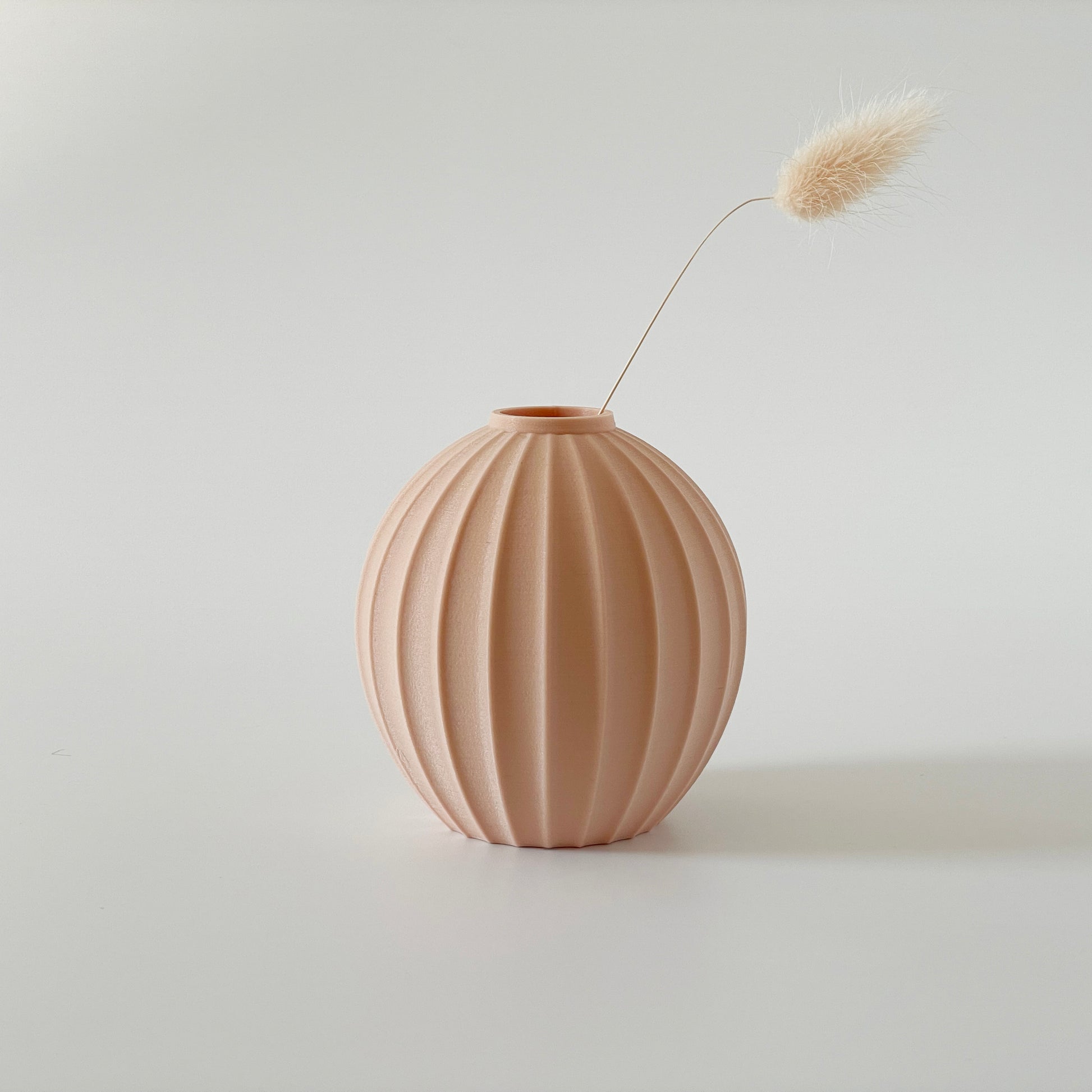 A beige spherical, 3D printed vase with a ribbed design, containing a dried plant, on a white background.