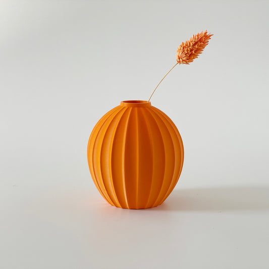 An orange 3D printed vase with a ribbed design, displaying a dried flower inside against a white background.