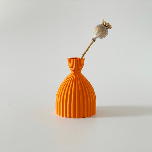 An orange 3D printed hour glass shaped vase with a ribbed design, displaying a dried flower inside against a white background.
