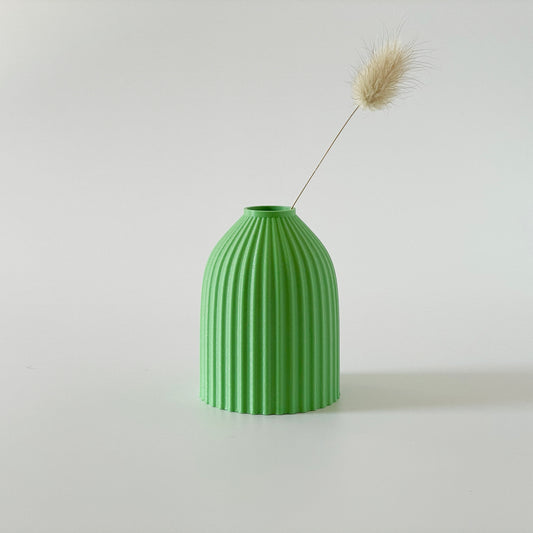 A green, 3D printed vase with a ribbed design, containing a sprig of dried grasses.
