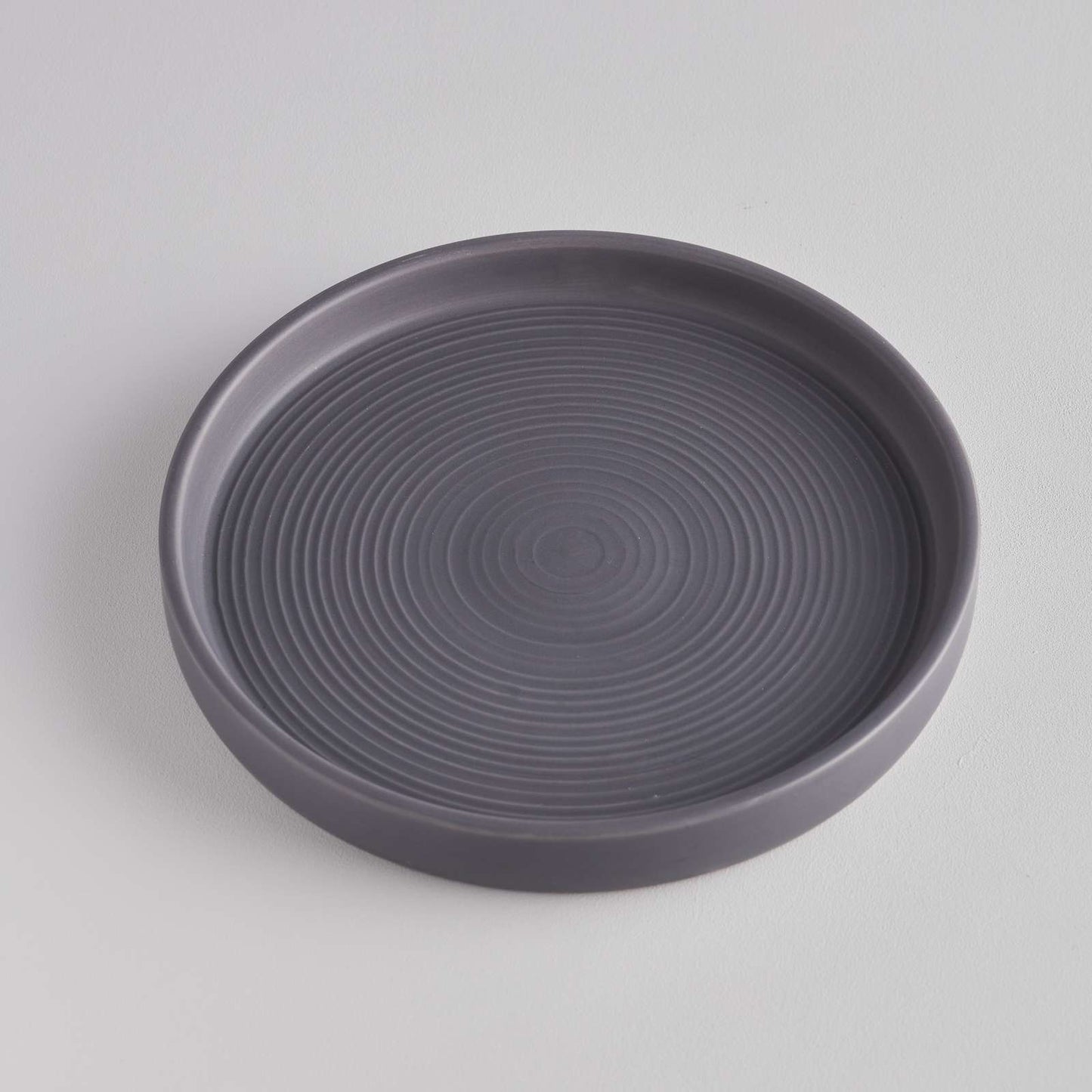 A simple, round grey dish with a smooth surface and concentric circular patterns etched into the base. The dish is set against a light grey background, highlighting its minimalist design and subtle texture.