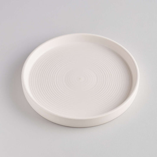 A simple, round white ceramic dish with a smooth surface and subtle concentric circular patterns. The dish is set against a light grey background, highlighting its minimalist design and clean lines.