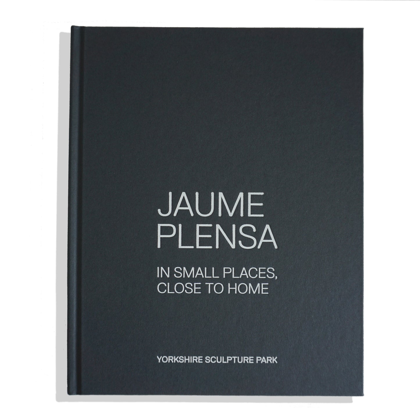 A closed book titled "Jaume Plensa: In Small Places, Close to Home" with a dark green, blue cover and white text. The book is associated with the Yorkshire Sculpture Park, suggesting a focus on the artist's work and themes.
