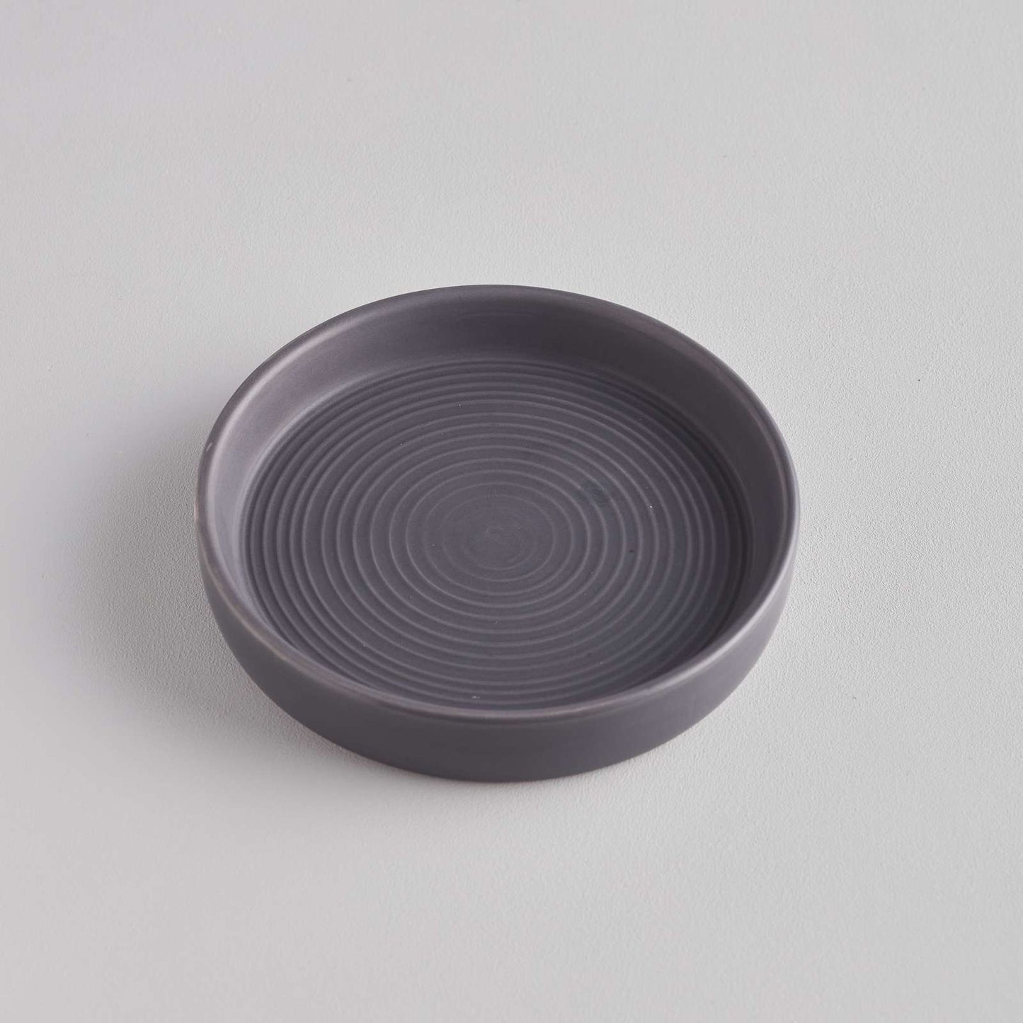 A simple, round grey dish with a smooth surface and concentric circular patterns etched into the base. The dish is set against a light grey background, highlighting its minimalist design and subtle texture.