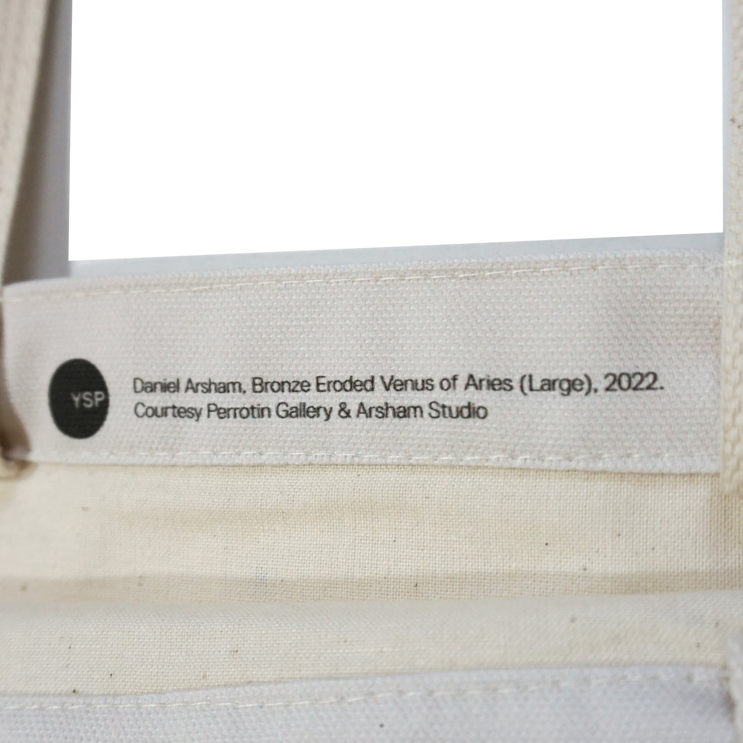 A close-up of a label attached to a canvas bag, featuring the text: "Daniel Arsham, Bronze Eroded Venus of Aries (Large), 2022. Courtesy Perrotin Gallery & Arsham Studio." The label is printed in black on a light background.