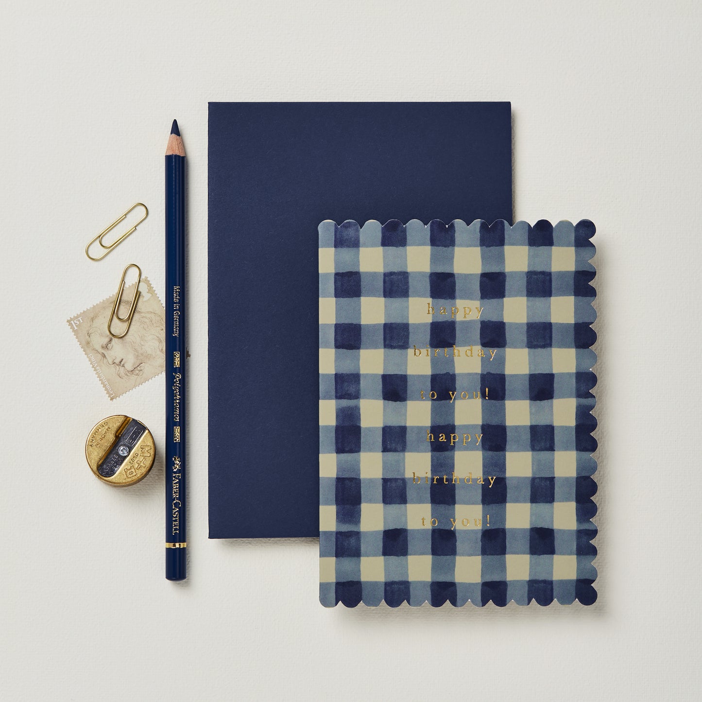 A flat lay of a navy blue birthday card with gold lettering, a matching envelope, a blue pencil, a round gold paperclip, a small piece of vintage paper, and a circular decorative item, all arranged on a light background.