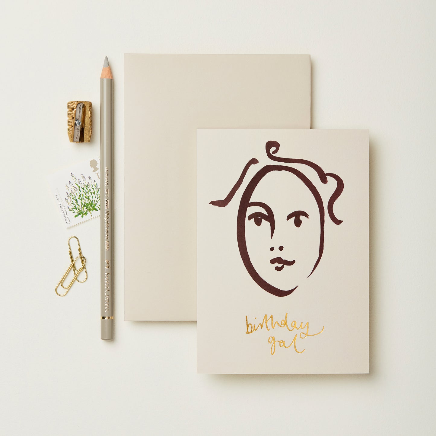 A flat lay featuring a minimalist birthday card with a line drawing of a face and the text "birthday gal" in gold. Beside it are a blank notepad, a grey pencil, a small sharpener, a paperclip, and a floral stamp on a light background.