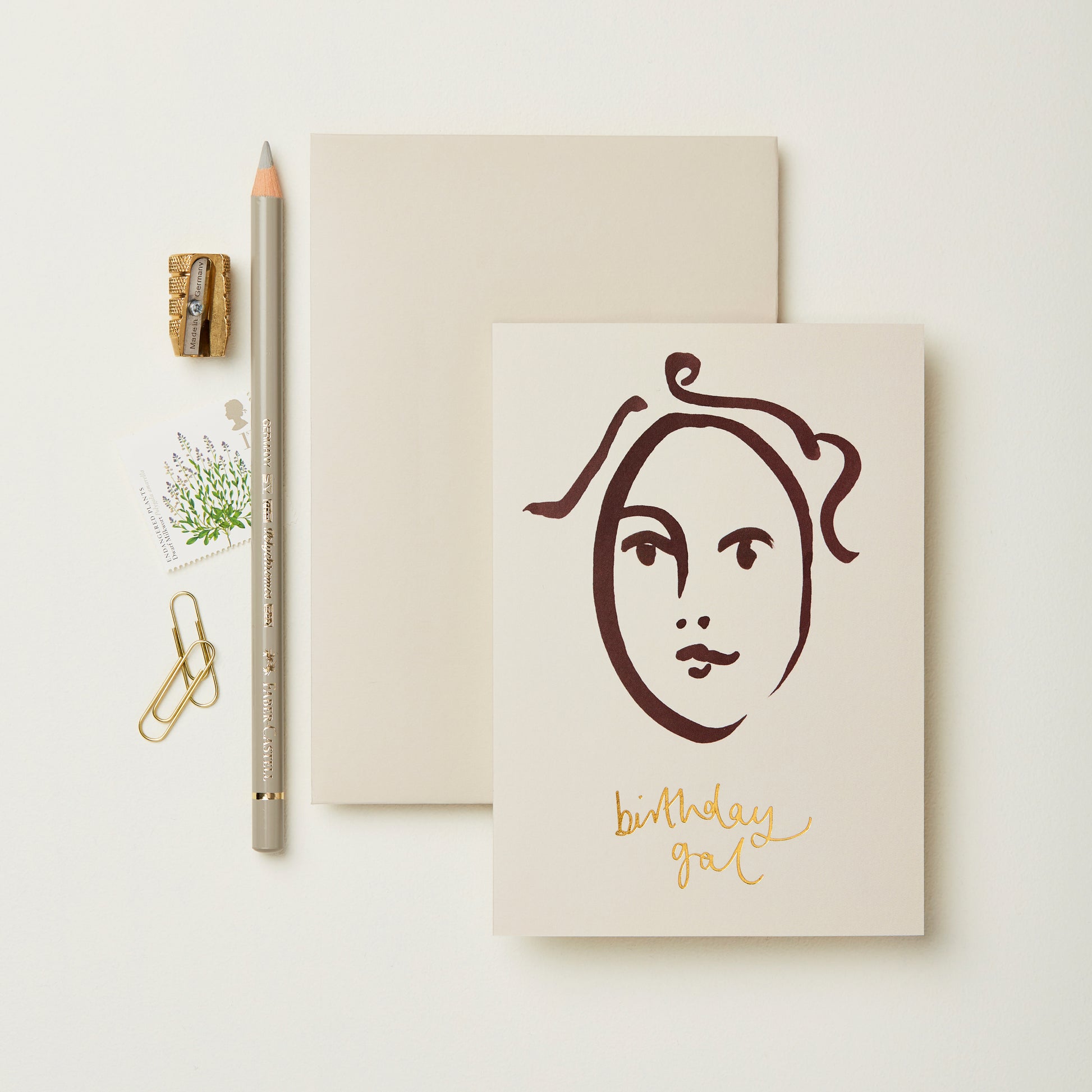 A flat lay featuring a minimalist birthday card with a line drawing of a face and the text "birthday gal" in gold. Beside it are a blank notepad, a grey pencil, a small sharpener, a paperclip, and a floral stamp on a light background.