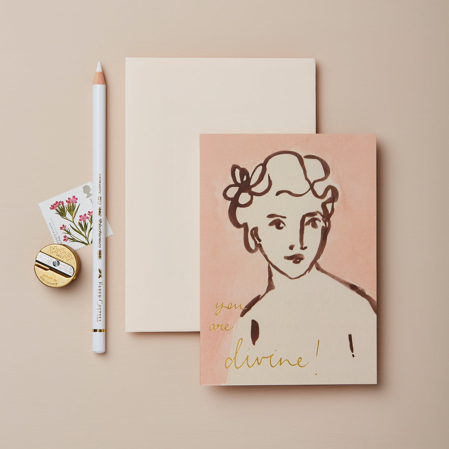 A flat lay featuring a greeting card with a minimalist illustration of a woman and the text "you are divine!" in gold. Beside it, a blank notepad, a white pencil, and a floral stamp on a beige background.