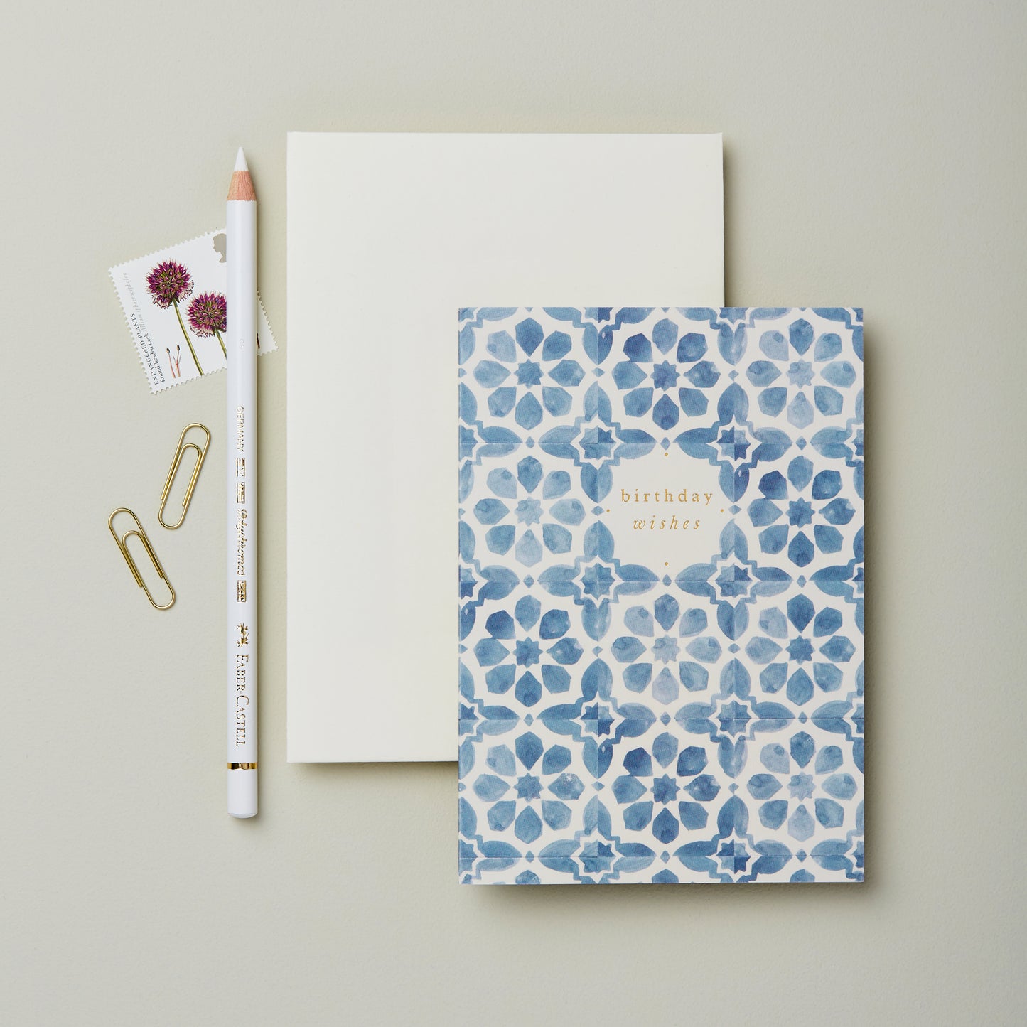 A flat lay featuring a birthday card with a blue floral pattern, a blank white notepad, a white pencil, two paper clips, and two stamps with floral designs, all arranged on a light grey background.