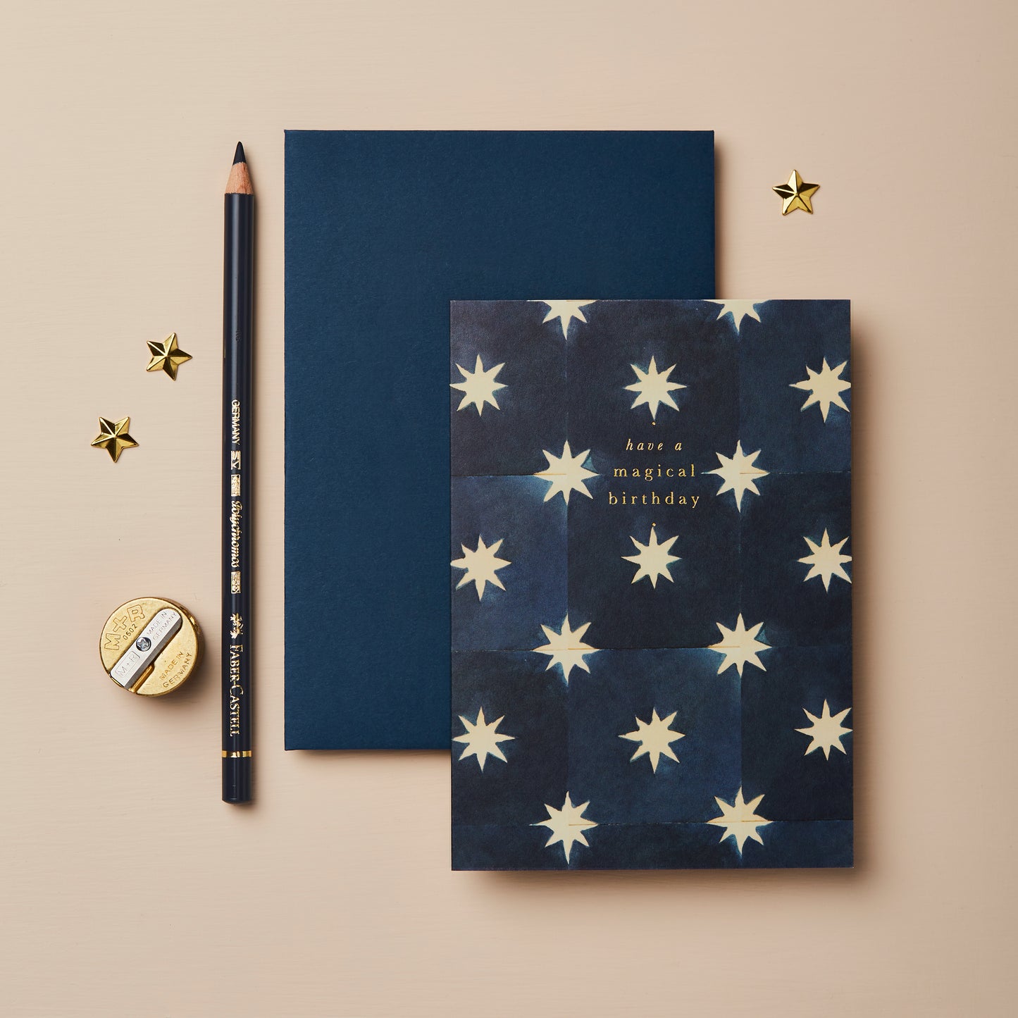 A flat lay featuring a dark blue birthday card adorned with cream stars, inscribed with "Have a magical birthday." Beside it, a black pencil, a navy blue envelope, and a gold sharpener, with a few small gold stars scattered around.