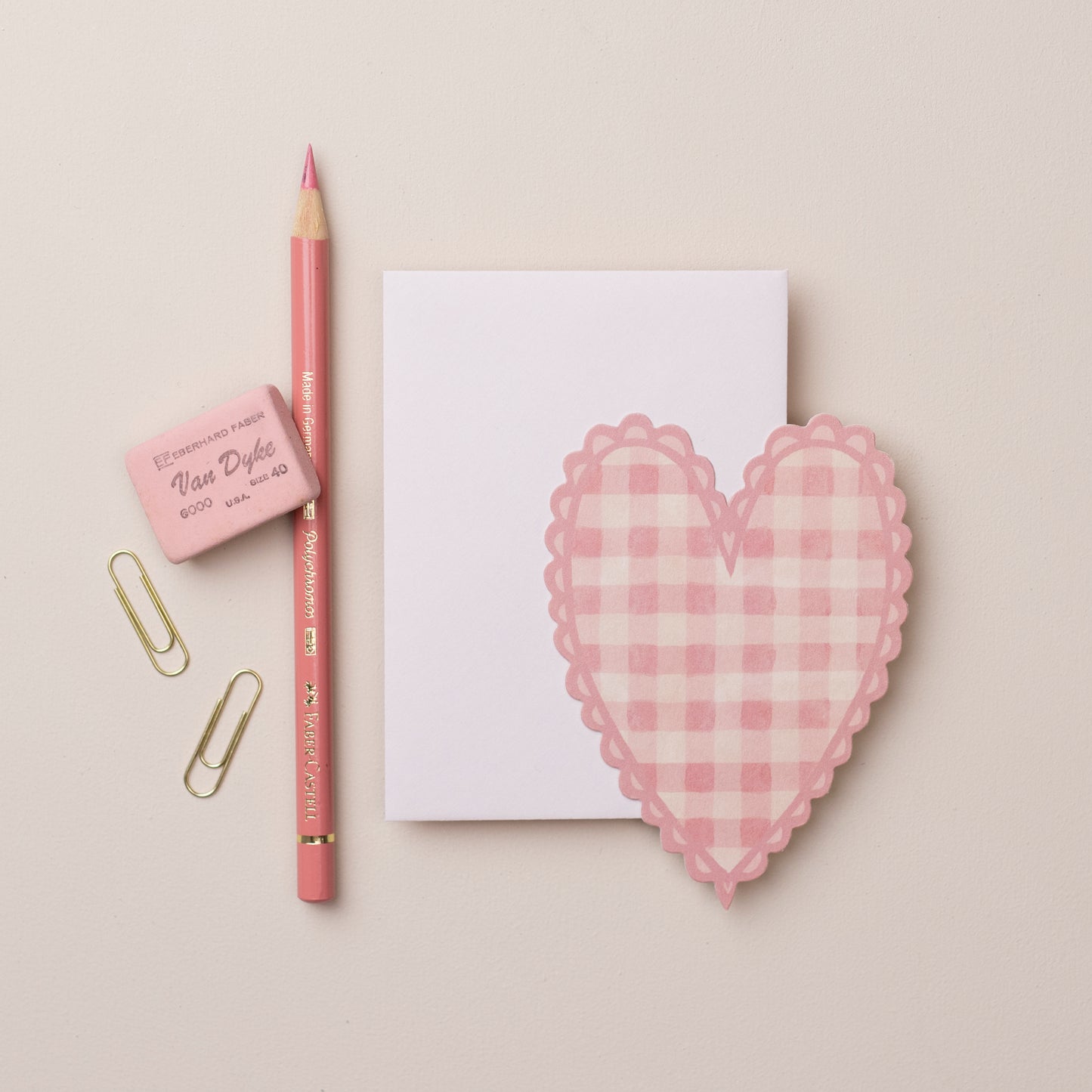 A flat lay featuring a blank notepad, a pink gingham heart-shaped mini card, a pink pencil, and two paper clips. The background is a soft beige, creating a warm and inviting aesthetic.