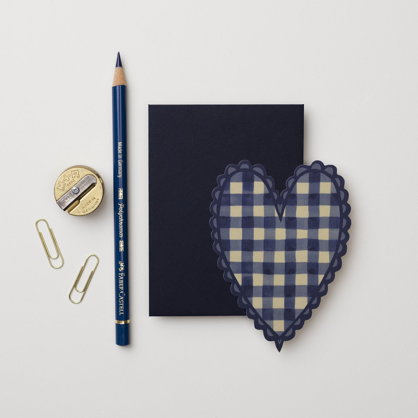 A flat lay featuring a navy blue notepad, a blue pencil, a gold pencil sharpner, two paper clips, and a heart-shaped mini card in beige and navy gingham. The arrangement is on a light background.