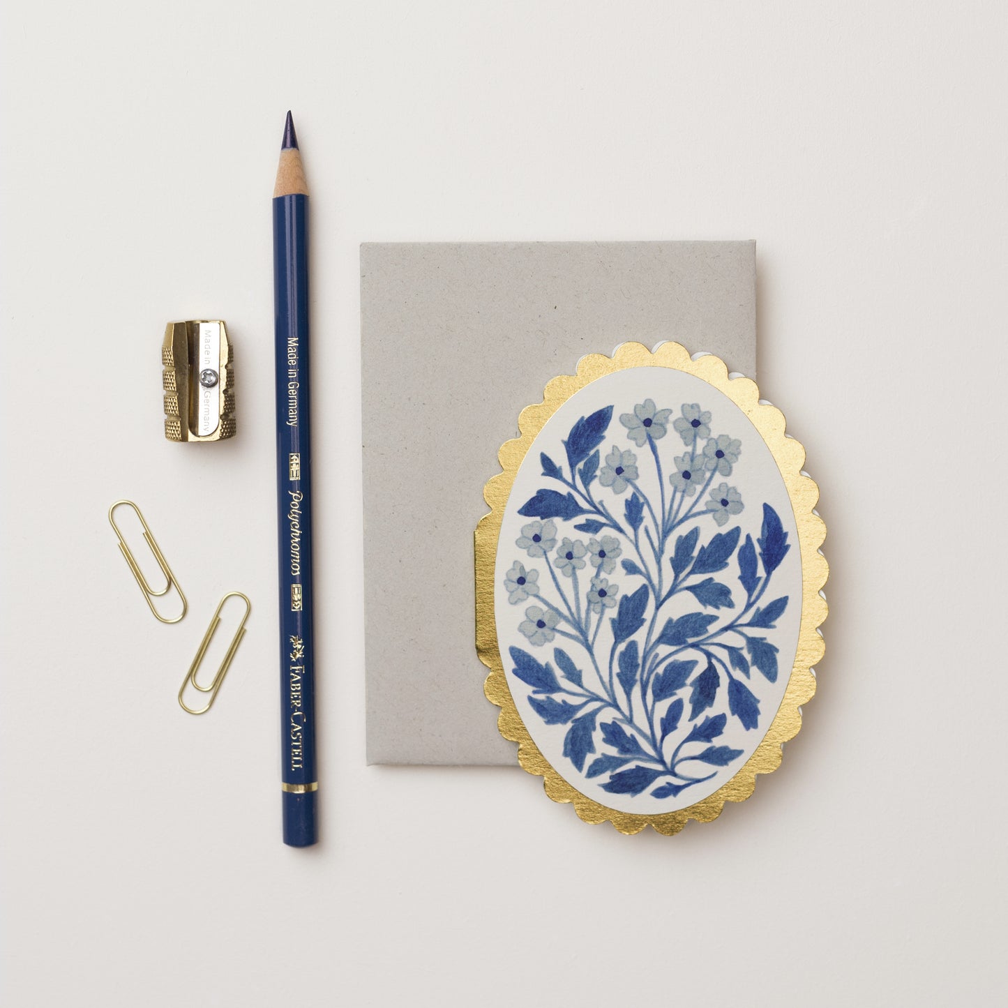A flat lay arrangement featuring a grey notepad, a blue pencil, a gold pencil sharpener, two gold paperclips, and an oval decorative mini card with a blue floral design on a white background, all set against a light surface.