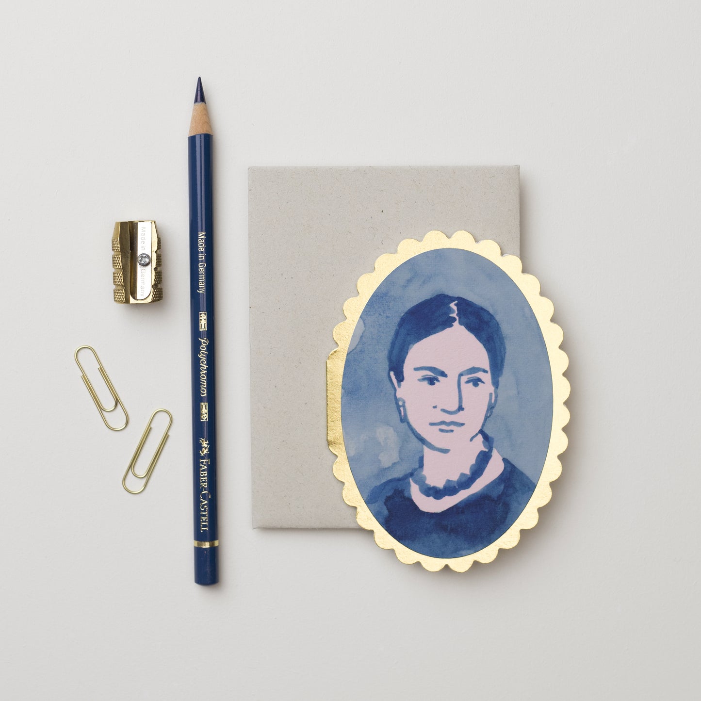A flat lay featuring a grey notepad, a gold-edged oval mini card featuring portrait of a woman with dark hair and a necklace, a blue pencil, a metal sharpener, and two paper clips. The background is a light, neutral colour.